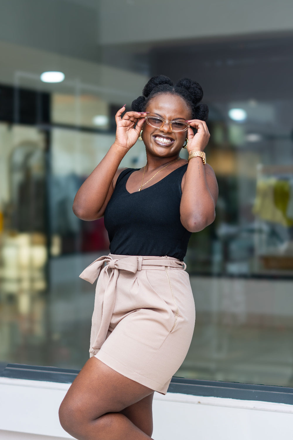 Woman wearing a black top and beige shorts, standing in front of a glass window.