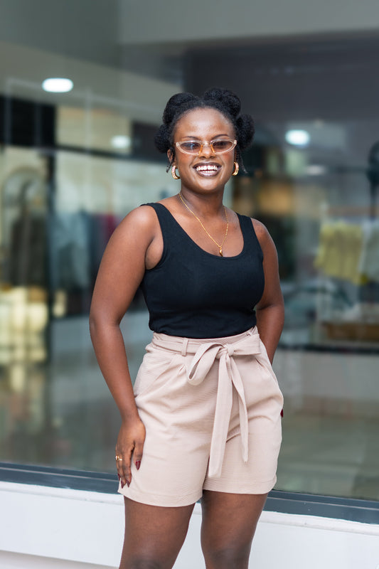 Woman wearing a black top and beige shorts with sunglasses, standing in front of a glass wall.
