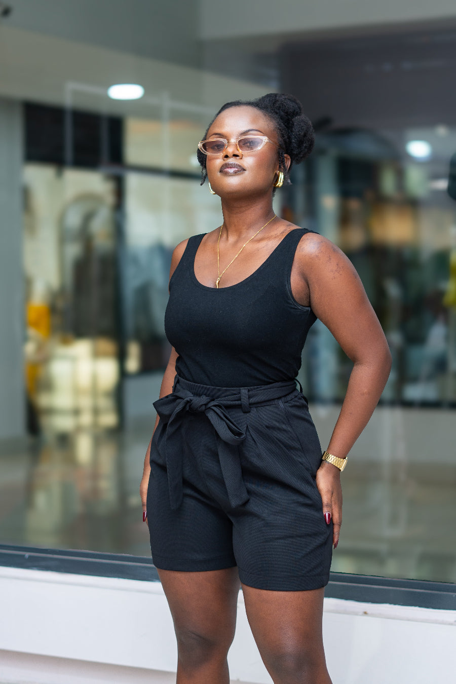 Woman wearing a black sleeveless top and shorts in front of a reflective glass wall.